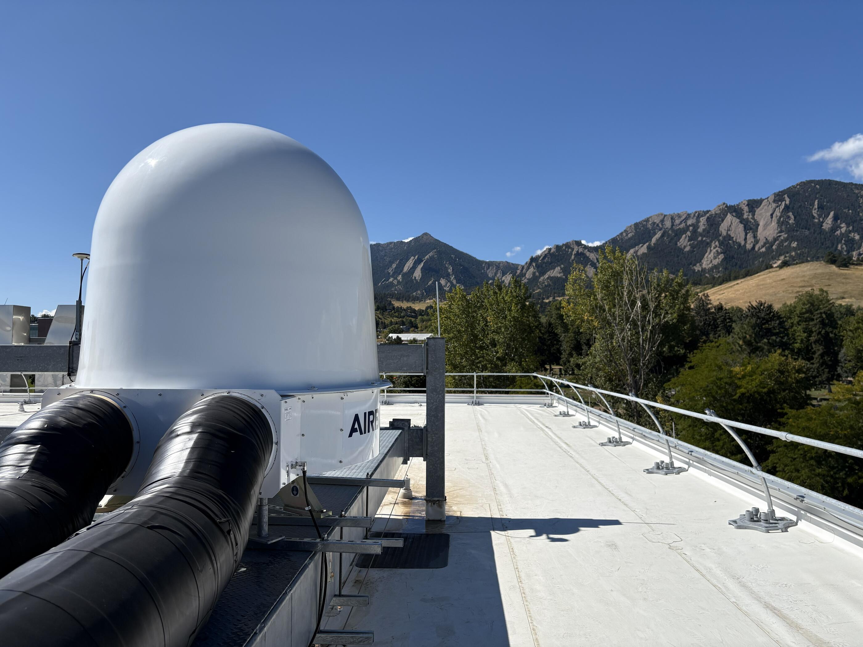 A white enclosure for a satellite antenna stands on a Boulder rooftop, with the Flatirons in the background.