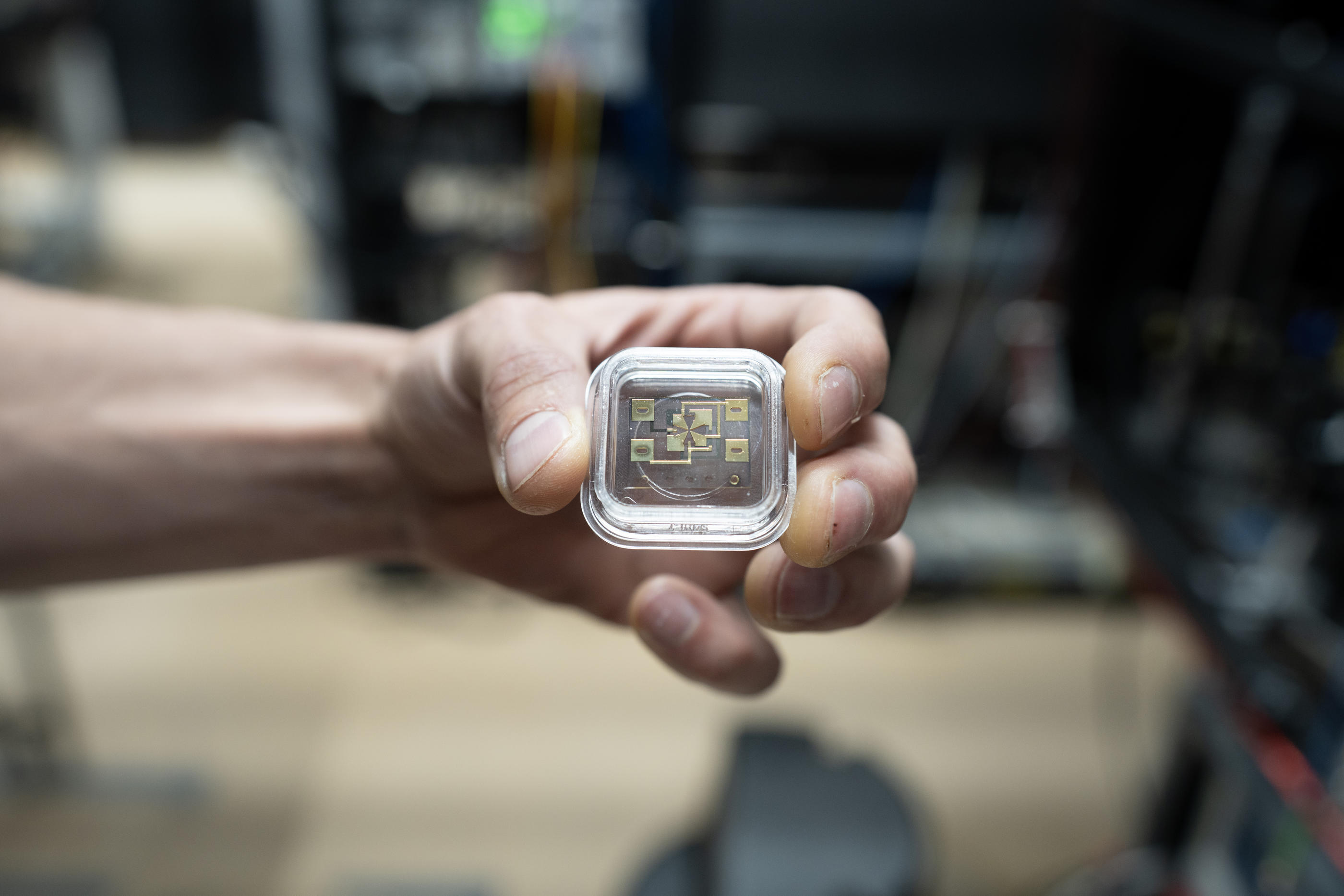 A hand holds a small clear case containing the gold-colored ion trap used in an atomic clock.