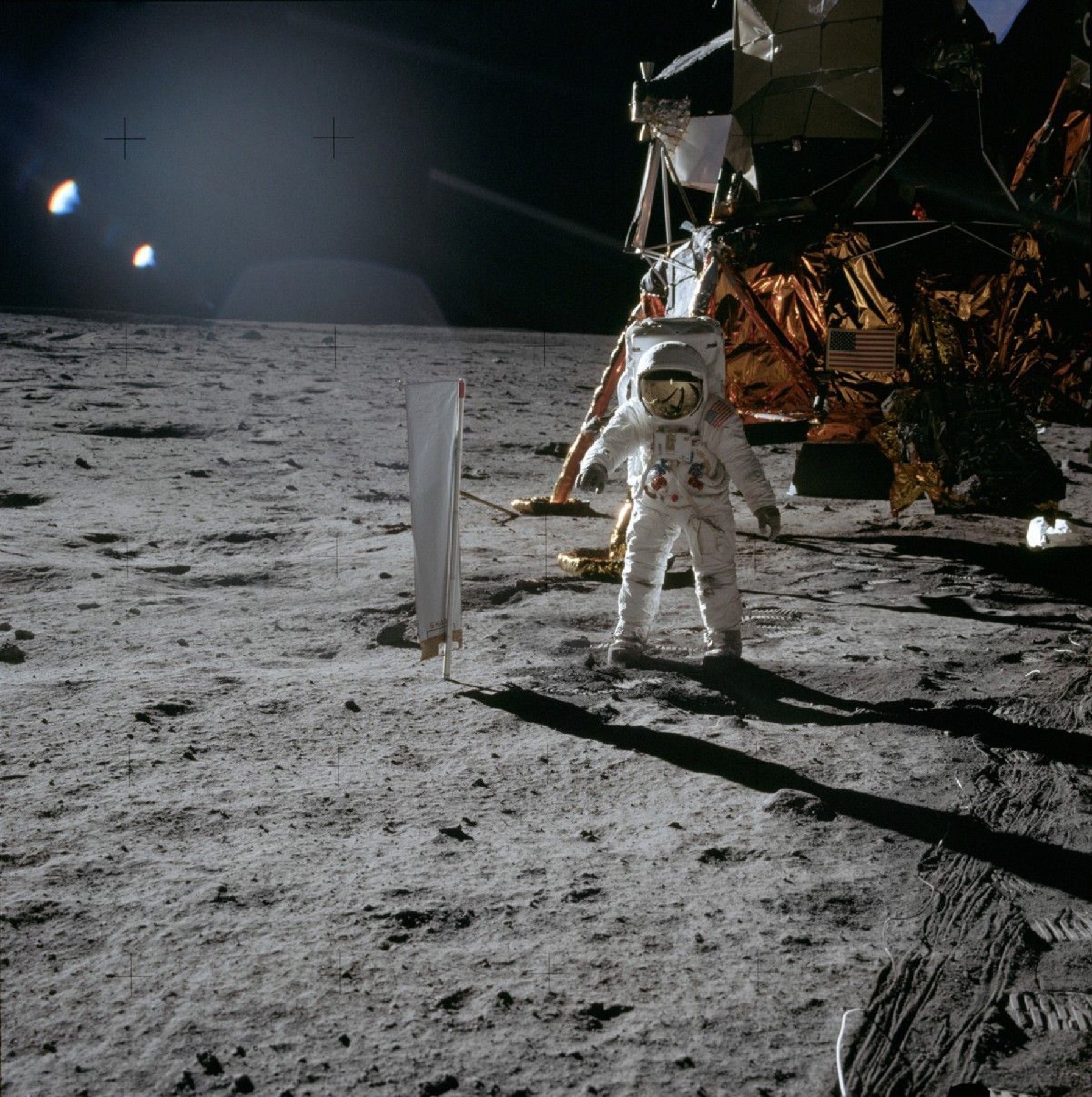 Buzz Aldrin stands beside a narrow upright sheet of foil on the lunar surface, with the lunar module behind him.