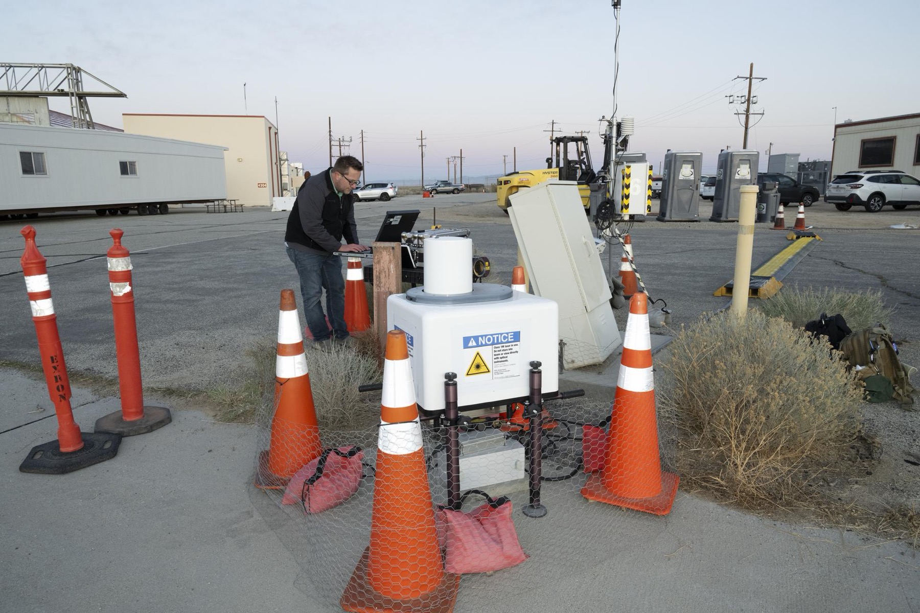 A researcher works at a laptop beside a white wind-lidar instrument surrounded by safety cones.
