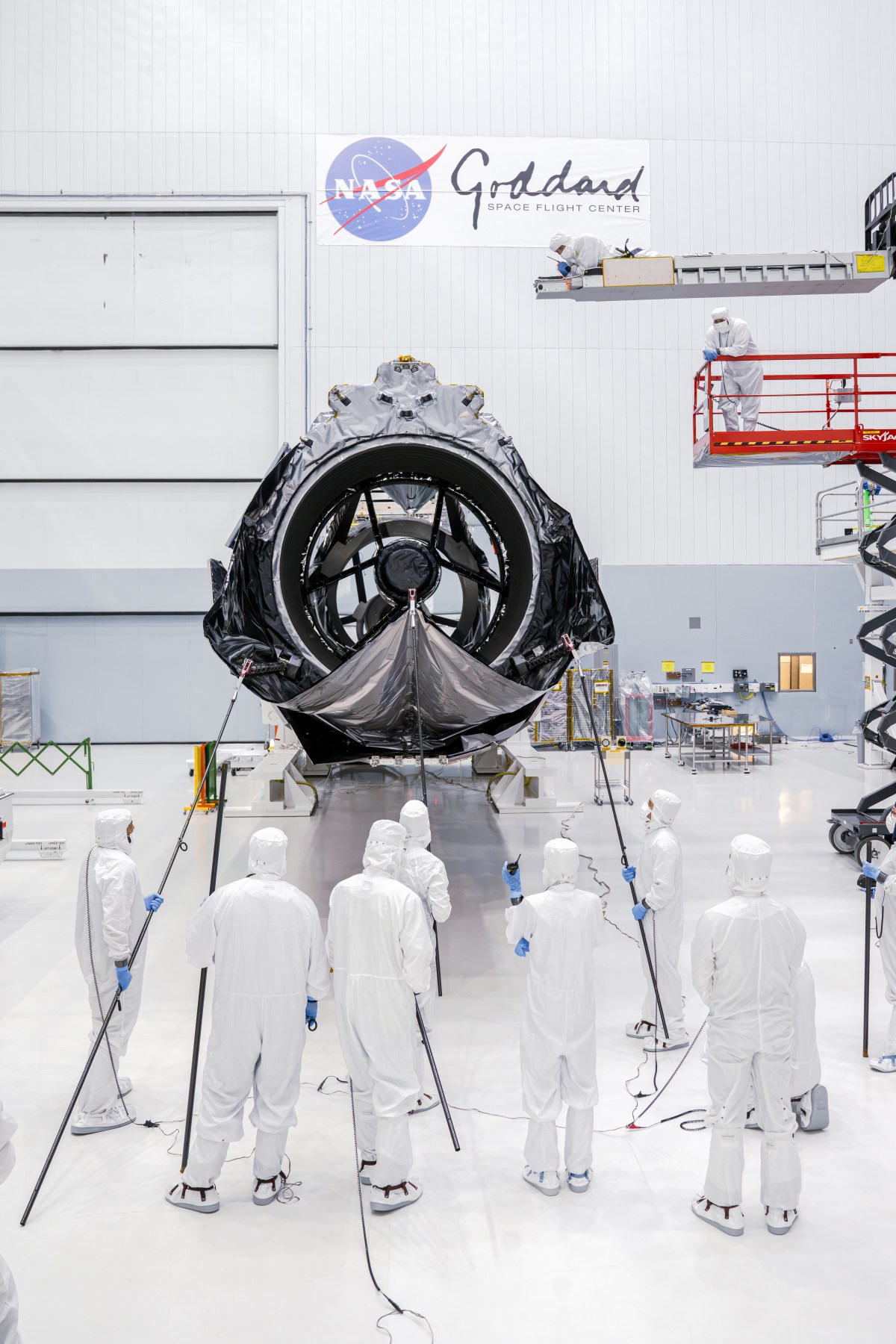 Technicians in clean-room clothing lower a flexible cover across the opening of Roman’s telescope.