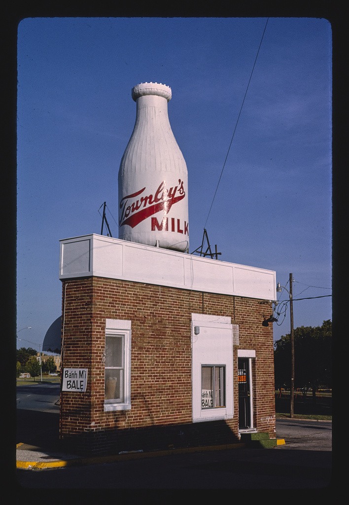A giant Townley’s Milk bottle rises above a small brick storefront under a blue sky.