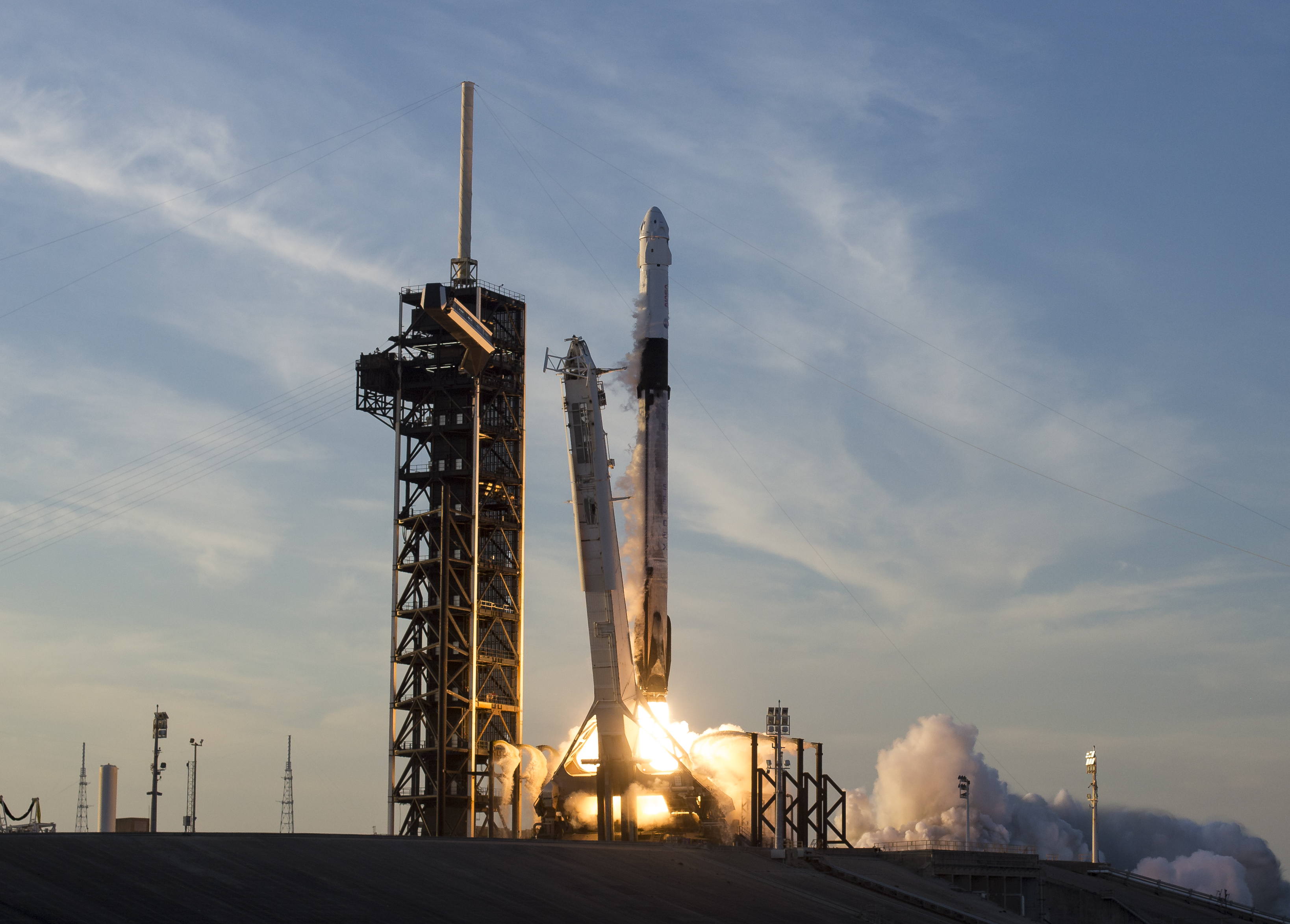 A Falcon 9 carrying a Dragon capsule lifts off beside its launch tower.