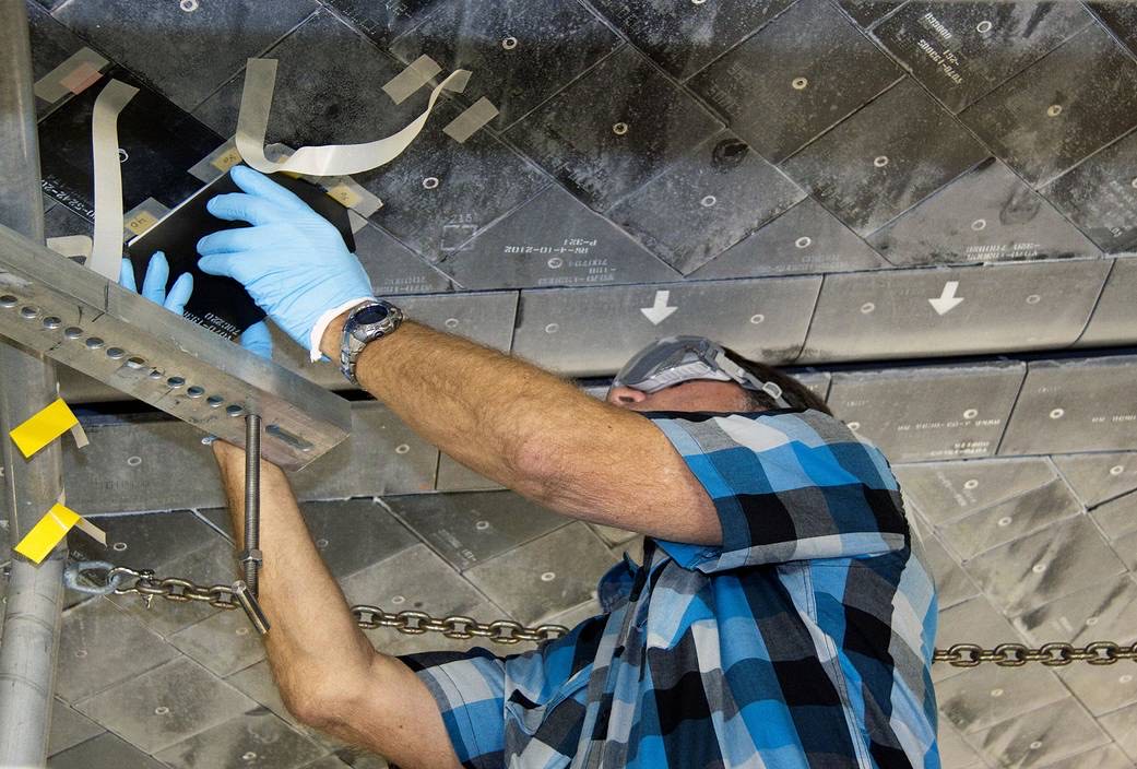 A technician wearing blue gloves checks a tile opening beneath space shuttle Atlantis, surrounded by numbered heat-shield tiles.