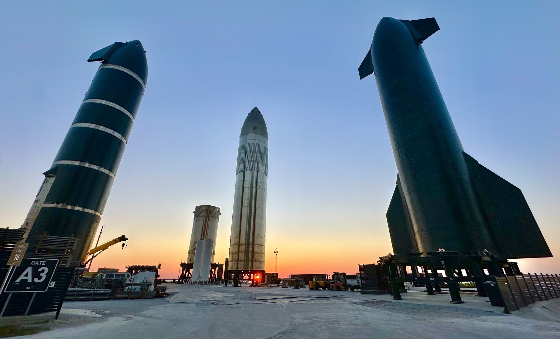 Several Starship prototypes and booster hardware stand in Starbase’s rocket garden against a blue and orange sunset.