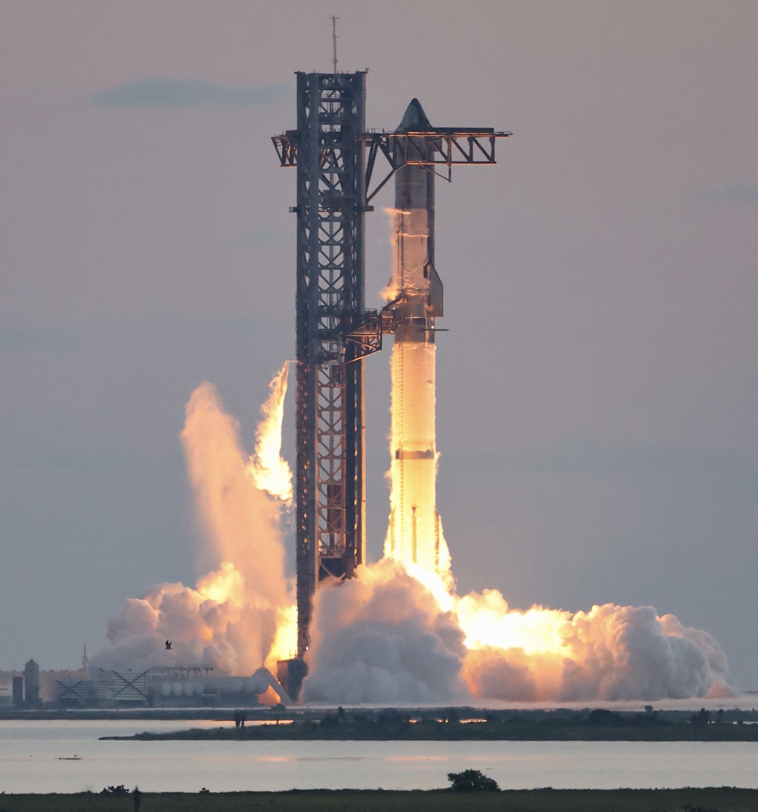 Starship beside its launch tower, with orange engine exhaust and clouds spreading across the pad.