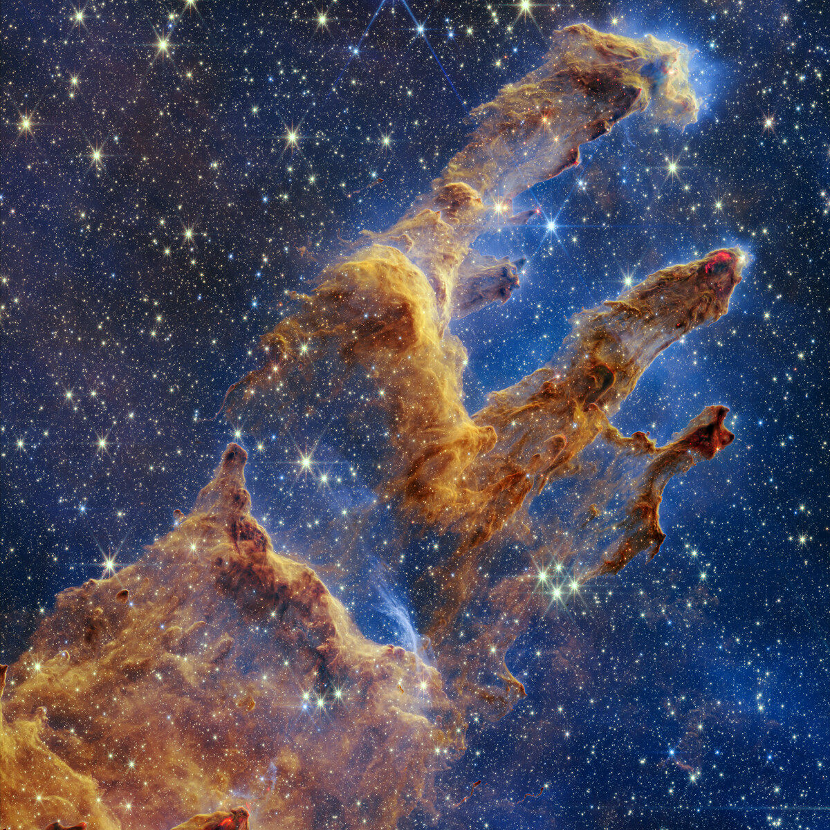 Webb's processed near-infrared observation of the Pillars of Creation, showing gold and brown gas-and-dust structures surrounded by stars against blue space.